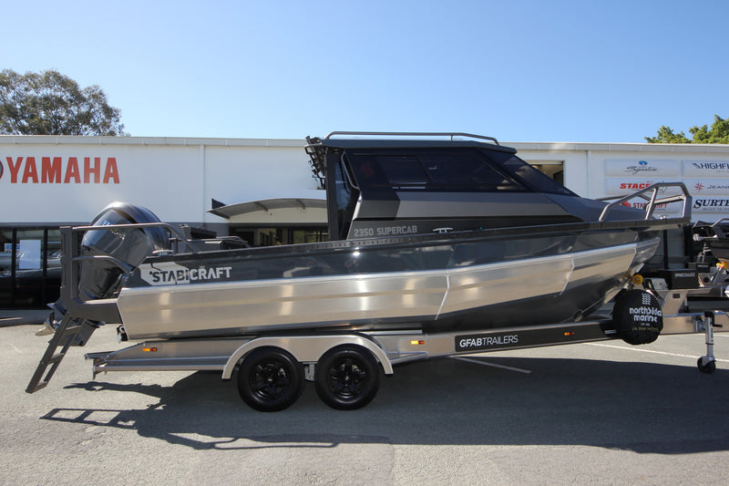 A Stabicraft Supercab boat with a dark gray and metallic silver hull, mounted on a dual-axle gray trailer, parked outdoors in front of a dealership building.