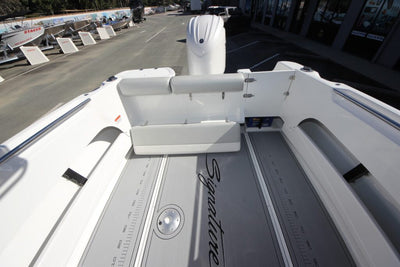 A high-angle view of the cockpit and deck of a white boat, showing a white dual-seat bench, a white outboard motor mount, gray measurement markings on the deck, and the word 'Signature' printed on the deck. The boat is docked in a marina with other boats and buildings visible in the background.